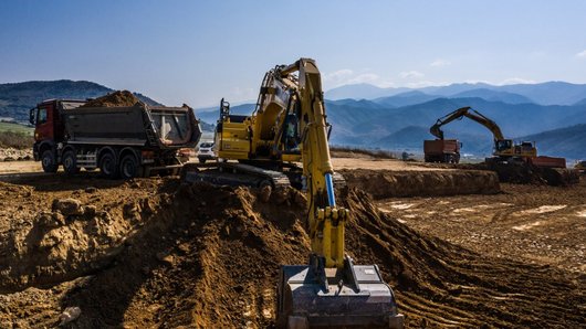 A construction site in a mountainous landscape where several excavators and a lorry are working. The background features mountains and blue sky.