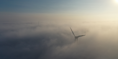 Wind turbines rise out of a dense blanket of fog, their tips visible above the mist.