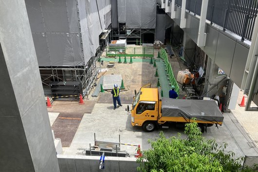 Eine Baustelle in einem engen Innenhof mit einem gelben Lastwagen, mehreren Arbeitern und grünen Absperrungen.
