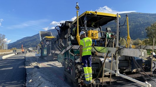 Straßenbauarbeiten, bei denen Bauarbeiter eine Asphaltiermaschine bedienen.