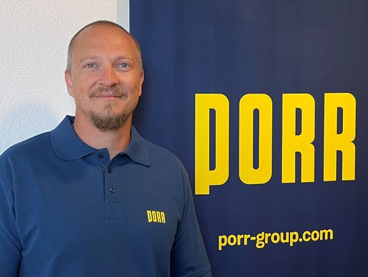 Ein Mann in einem blauen Polohemd mit PORR-Logo steht vor einem blauen Banner.