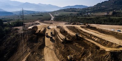 A construction site in a hilly landscape with several excavators and lorries at work. A road extends in the background, surrounded by mountains.