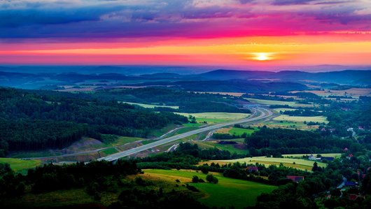 Landschaft mit Wiesen und Wäldern, durch die eine Straße führt, Sonnenuntergang im Hintergrund