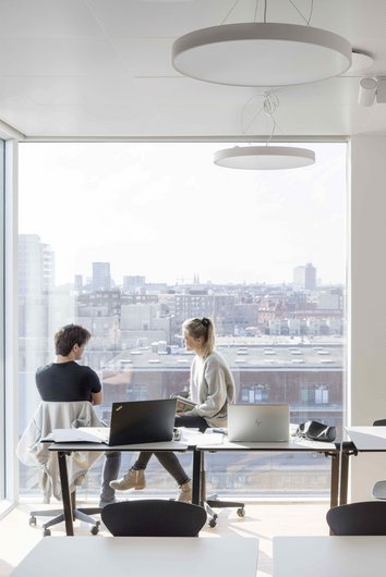 Auf dem Foto sind zwei Personen in einem hellen Büro mit großen Fenstern und Blick auf eine Stadtlandschaft zu sehen. Sie sitzen an einem Tisch mit Laptops und unterhalten sich, während sie die Aussicht genießen.