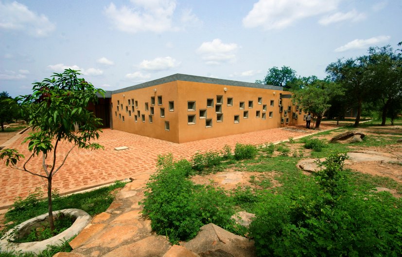 A modern building with a flat roof construction and many small windows, surrounded by paved areas and green vegetation.