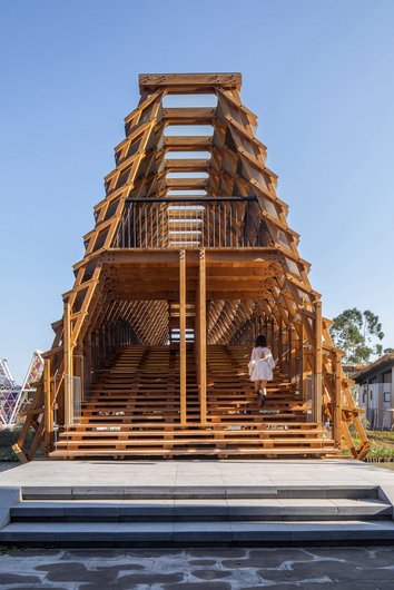 A modern wooden bridge with an open structure and stairs leading to an upper level where a person dressed in white is walking.