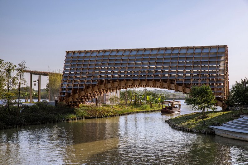 A wooden arch with a lattice structure spans a calm river and connects two riverside landscapes.