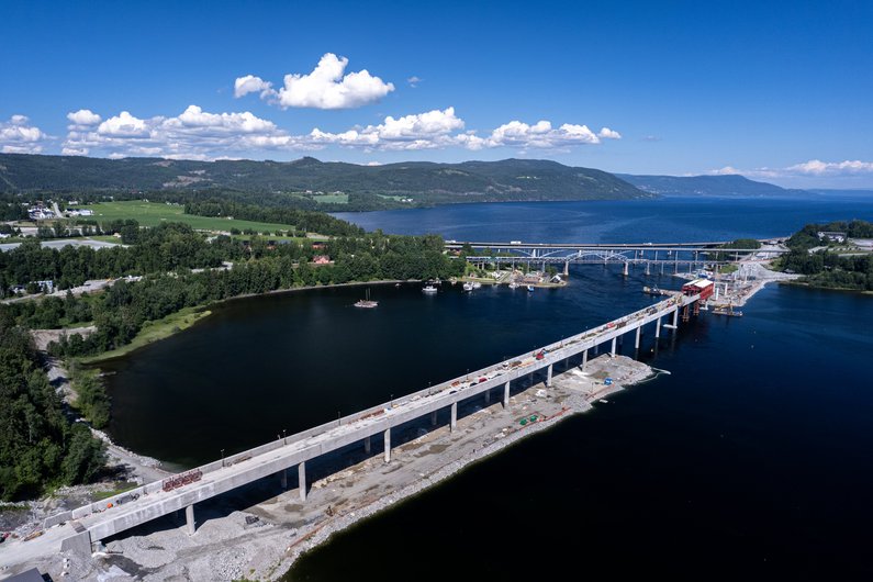 Eine im Bau befindliche Brücke über einen großen Fluss oder See, umgeben von Wäldern und Hügeln.