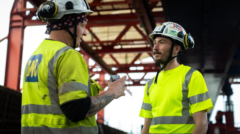 Two construction workers in yellow safety waistcoats and hard hats, standing on a building site and chatting.