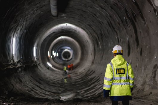 Frau mit Helm und gelber Jacke von hinten vor einer Tunnelröhre