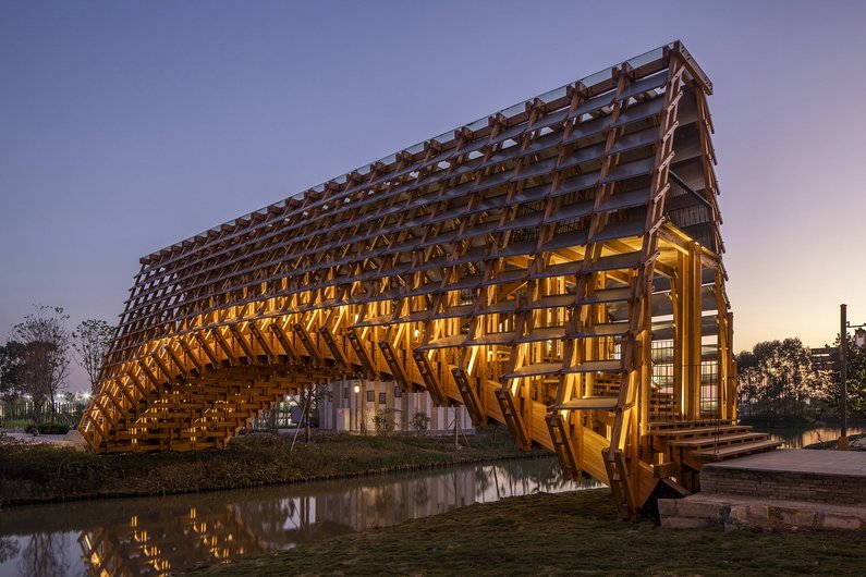 An illuminated wooden bridge with an open lattice structure that crosses a small river.