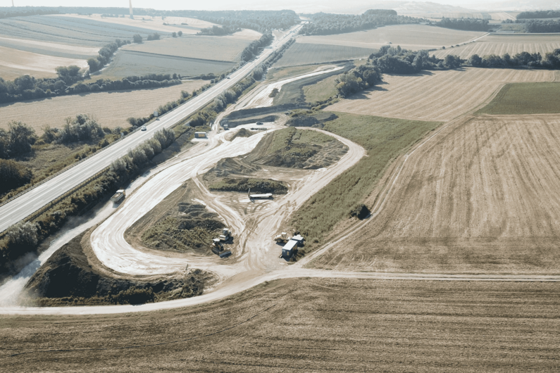The photo shows an aerial view of a construction site next to a country road, surrounded by wide fields and a few trees. The construction site includes several gravel paths and construction equipment, while the road runs through the rural landscape.