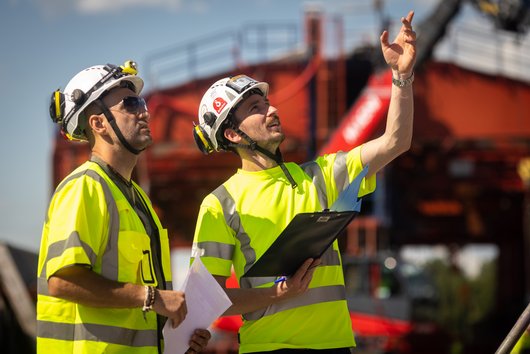 Two construction workers in yellow safety waistcoats and hard hats standing on a building site.