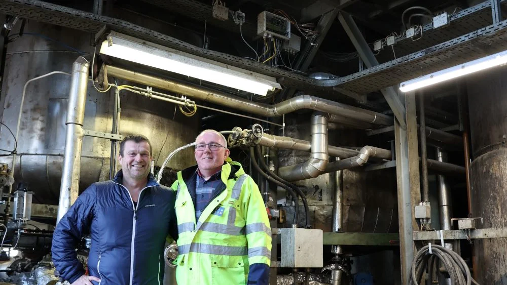 Zwei MÀnner stehen vor Stahlkesseln und lÀcheln, der linke trÀgt eine baue Jacke und hat seinen Arm um die Schultern des anderen gelegt