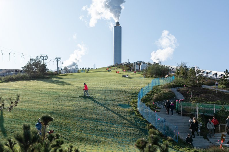 People skiing down an artificial, green ski slope, a smoking chimney can be seen in the background.