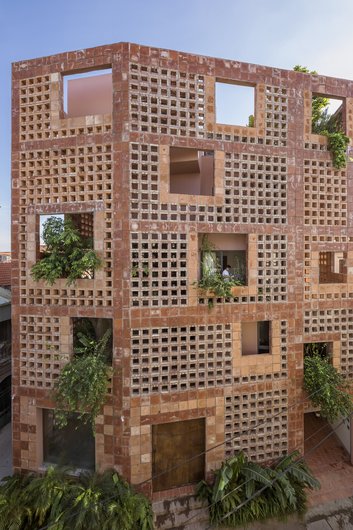 A modern building with a striking red brick façade and large, open windows. It is partially surrounded by plants and greenery.