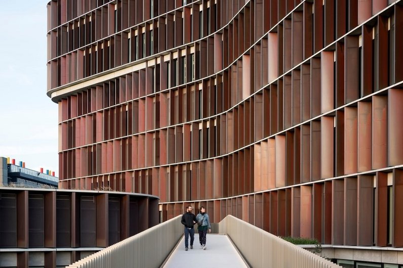 Two people walk in front of a modern building with a wave-shaped, reddish-brown façade.