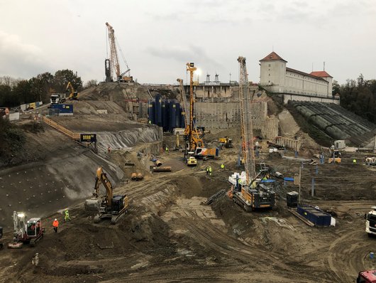 Eine große Baustelle mit mehreren Baugeräten und Arbeitern, die an einer tiefen Baugrube arbeiten.