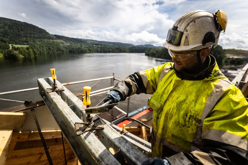 A construction worker adjusts a tool on scaffolding on a riverbank while working in full protective clothing and helmet.