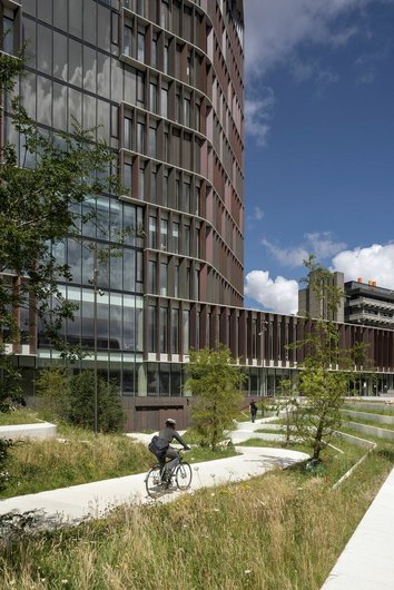 Modernes Gebäude mit geschwungener Fassade, Radfahrer auf einem Weg durch grüne Wiesen im Vordergrund.