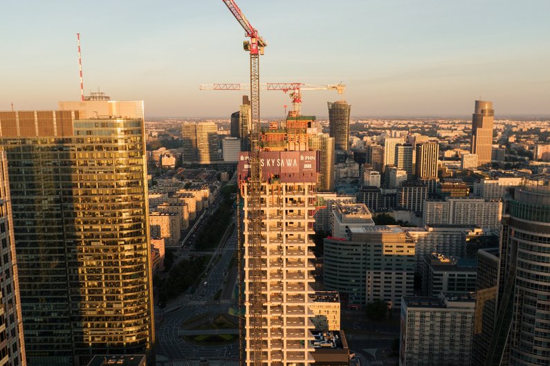 A cityscape at sunset with a tall building under construction and a crane on the roof, surrounded by finished skyscrapers and other construction cranes in the background.