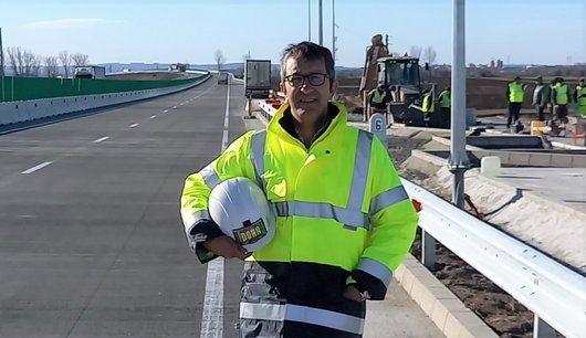 A smiling construction worker in a yellow safety jacket and helmet standing on a newly built road.