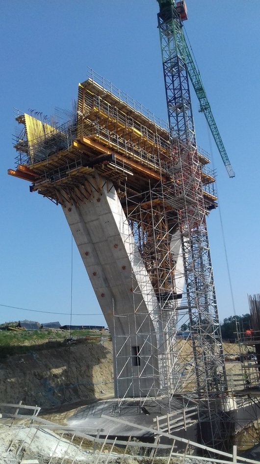 Eine im Bau befindliche Brückenstütze mit einem komplexen Gerüst und einem Kran, umgeben von einer Baustelle.