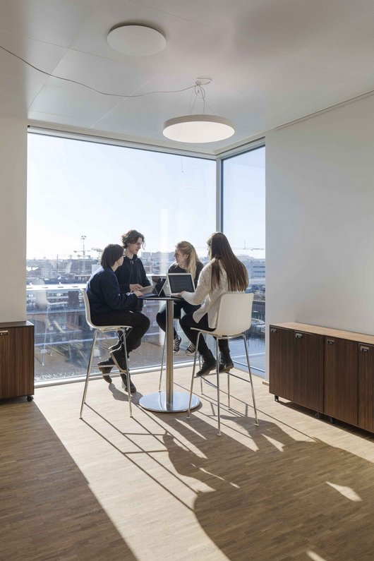 Das Foto zeigt vier Personen, die an einem hohen Tisch mit Barhockern in einem modernen Büro sitzen und miteinander sprechen. Im Hintergrund ist durch große Fenster eine städtische Landschaft bei Tageslicht zu sehen.