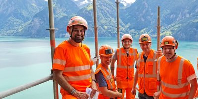 Five construction workers in orange-coloured protective clothing on scaffolding in front of an alpine scenery.