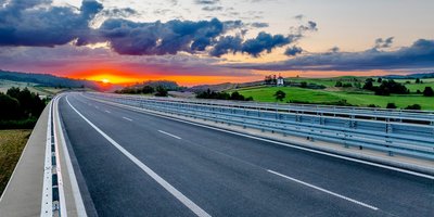 Straße mir Leitschienen, die durch eine grüne Landschaft führt mit einem Sonnenuntergang im Hintergrund