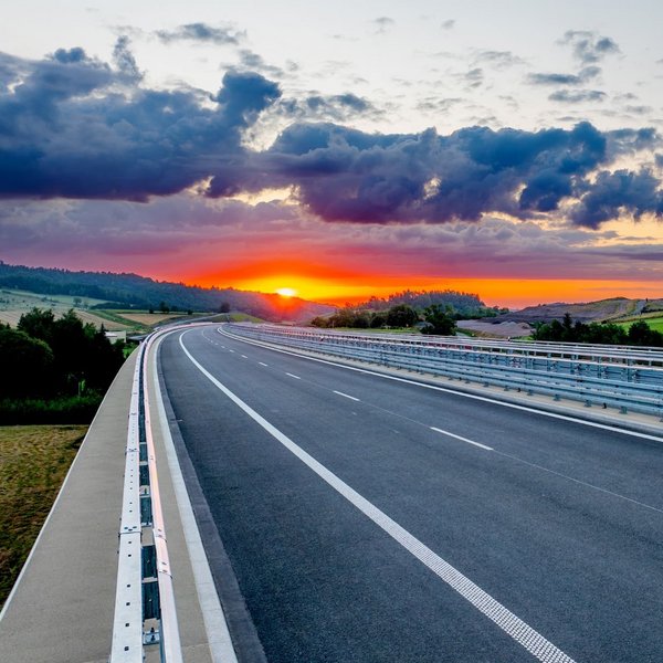 Straße mir Leitschienen, die durch eine grüne Landschaft führt mit einem Sonnenuntergang im Hintergrund