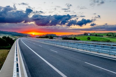 Straße mir Leitschienen, die durch eine grüne Landschaft führt mit einem Sonnenuntergang im Hintergrund