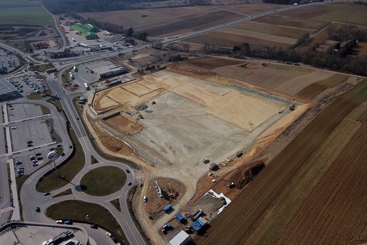 The photo shows an aerial view of a large construction site surrounded by fields and roads. In the foreground are a roundabout and a car park, while agricultural land and commercial buildings can be seen in the background.