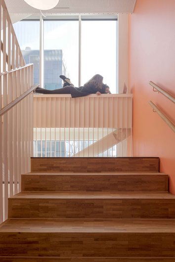 Auf dem Foto ist ein modernes Treppenhaus mit orangefarbenen Wänden und Holzstufen zu sehen. Im oberen Bereich der Treppe liegen zwei Personen auf einer Brüstung und arbeiten an einem Laptop, während sie durch ein großes Fenster Tageslicht genießen.