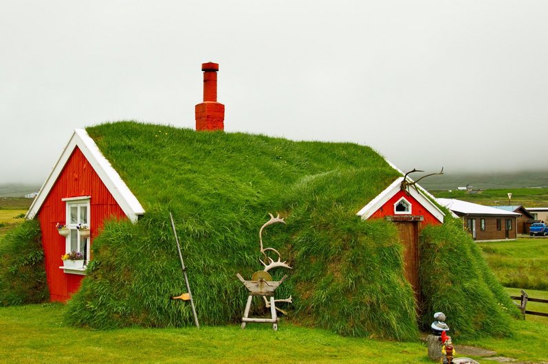 A traditional sod house with a red façade and grass-covered roof. A red chimney sticks out of the roof and rustic decorations stand in front of it.