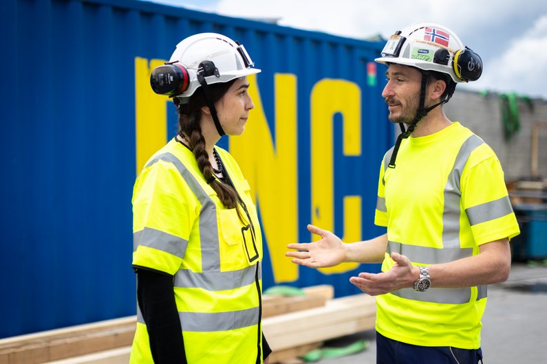 Two construction workers in yellow safety waistcoats and helmets, standing on a building site and talking to each other.