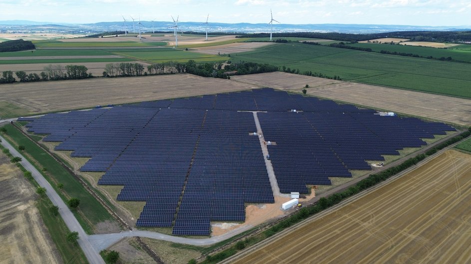 A large-scale solar park with numerous solar modules stretches across a field, with several wind turbines visible in the background.